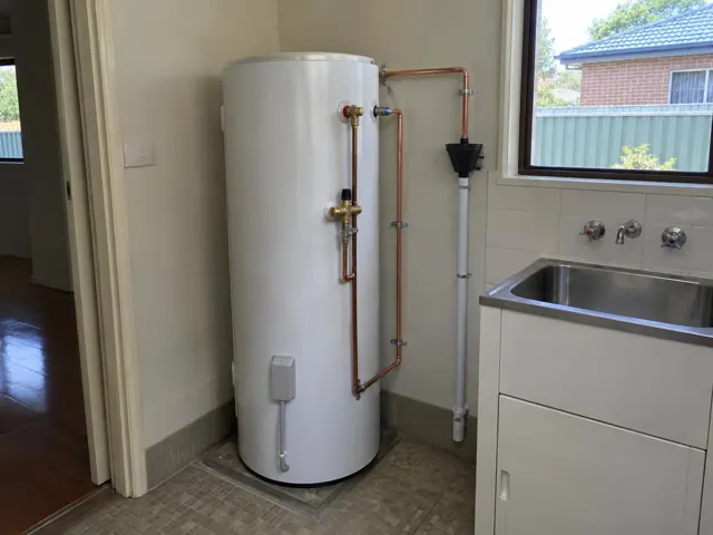 White hot water cylinder beside a laundry sink with copper and PVC pipework in a tiled room in Cessnock